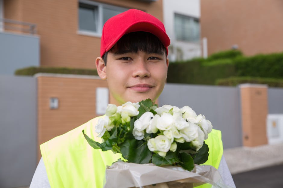 A young delivery person with a friendly expression, wearing a red cap and a high-visibility yellow vest, holding a bouquet of fresh white flowers wrapped in beige paper. The bouquet includes white ros