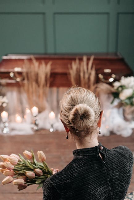 A woman with platinum blonde hair styled in a neat bun, seen from behind, holds a bouquet of pale pink tulips with green stems. She is dressed in a textured dark grey jacket with a small part of a bla