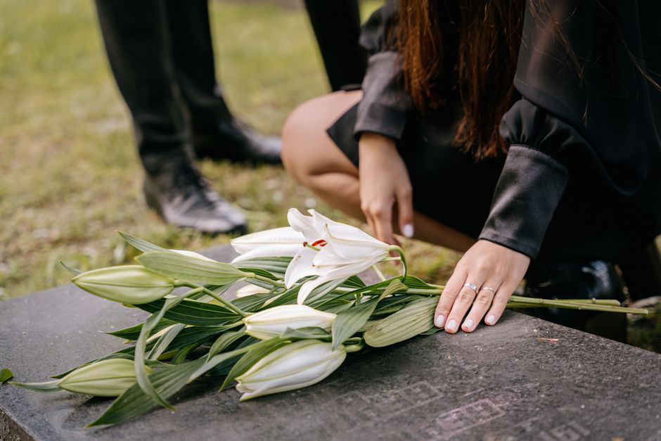 A person dressed in black is kneeling on a stone memorial or grave, gently holding a bouquet of white lilies with long green stems and leaves, which are freshly cut and perfectly arranged. The bouquet