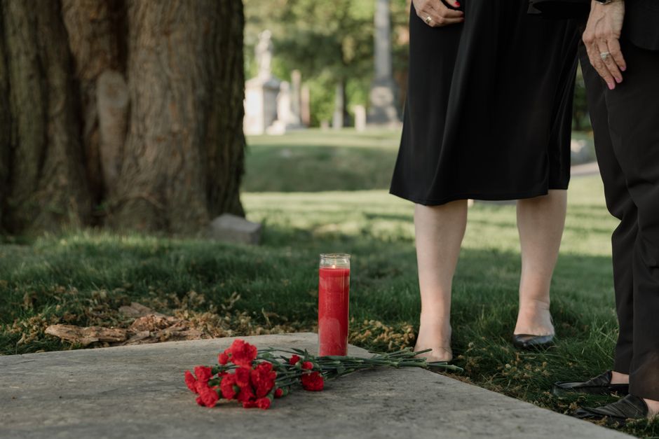 A floral memorial arrangement features a bouquet of vibrant red carnations placed on the ground beside a tall, clear glass candle holder filled with red wax. Partially visible are two individuals dres