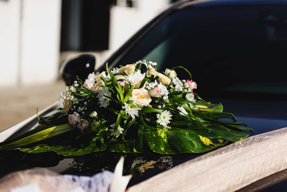 A beautifully arranged floral bouquet placed on the hood of a black car, featuring white and pale pink roses, white daisies, and delicate baby's breath, accented with lush green foliage and large glos
